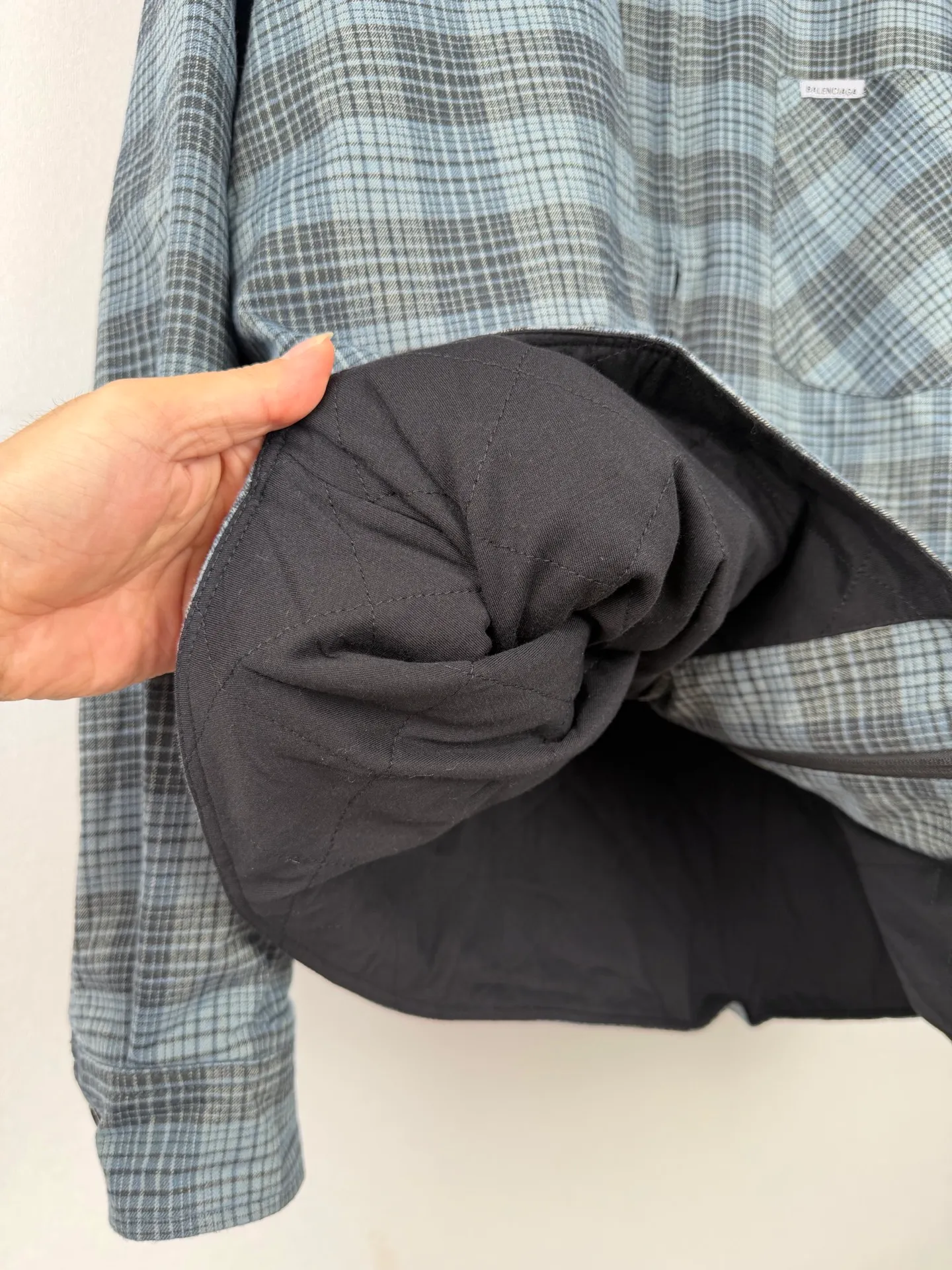 BALENCIAGA/balenciaga blue and gray plaid zipper pocket logo quilted jacket combines a three-layer structure of plaid woven fabric, filling cotton, and lining, and integrates plaid alignment technology and quilted garment production standards. There are multiple process difficulties in the whole process. The surface layer of the body is made of blue-gray Retro plaid fabric, and the cutting machine production strictly follows the global grid standards. The arrangement direction of the cutting pieces is fixed and cannot be changed at will. The horizontal and vertical lines of shoulder seams, side seams, and zipper placket joints must be consistent and coherent. The left chest patch pocket is cut independently, and the bag check pattern must be aligned with the front reference pattern. Slight deviations will destroy the visual integrity and directly increase fabric loss and cutting time. The most difficult part of the style is the three-layer composite sewing. The outer plaid fabric, filling cotton, and lining fabric have different physical properties, and the preshrinking processing parameters need to be adjusted separately. Pressure and temperature control are crucial during lamination operations. Once the parameters are out of balance, it can easily cause fabric stretch and distortion, plaid dislocation, and wrinkles and bulges in garments. At the same time, it is necessary to reasonably plan the quilting line to avoid the visual beauty of the grid pattern from being cut by the lines on the basis of firmly fixing the cotton layer and preventing cotton from running away. The placket adopts a centered zipper structure. It is difficult to sew the zipper tape and the quilted panels. It is required that the width of the left and right plackets be equal, the zipper routing is straight and smooth, and there is no lag in opening and closing. When the shirt lapel is filled with cotton, the thickness of the cotton needs to be controlled to prevent the collar from becoming bloated and unnaturally folded. The metal nameplate of the chest pocket is sewn at fixed points to avoid pulling the fabric during the operation and ensure that the plaid pattern is smooth and not deformed. The quilted material increases the overall thickness of the fabric. When sewing, you need to continuously adjust the presser foot pressure and stitch length to adapt to changes in sandwich thickness. The low-temperature and gentle ironing process is used for post-finishing to prevent the plaid from fading and the cotton layer from clumping due to high temperatures. From the precise alignment of cutting pieces, three-layer fabric pretreatment, composite quilting to garment finalization inspection, the processes are interlocking and the error tolerance rate is extremely low, testing the accuracy of plate making, equipment control and turning technology. Size:XS S M L